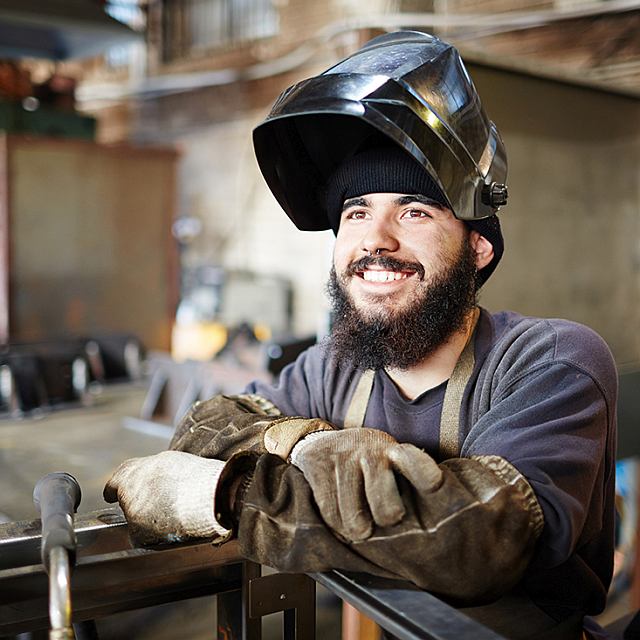 Junger Mann mit schmutzigen Ärmeln, der eine Schweißermaske trägt und sich in einer Fabrikhalle auf ein Metallteil stützt