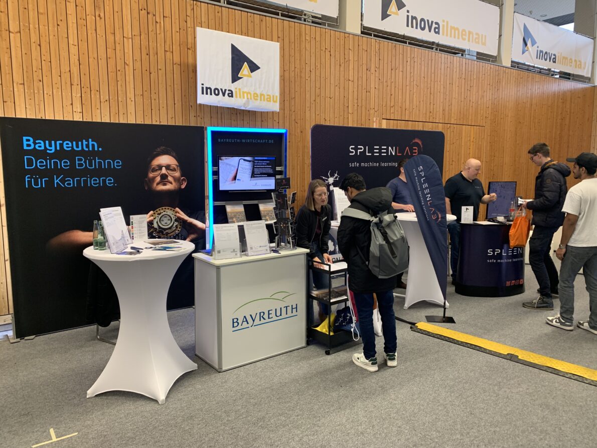 Eine Person steht an einem Messestand in einer Halle im Gespräch mit einer Frau. Im Hintergrund steht. Bayreuth. Deine Bühne für Karriere