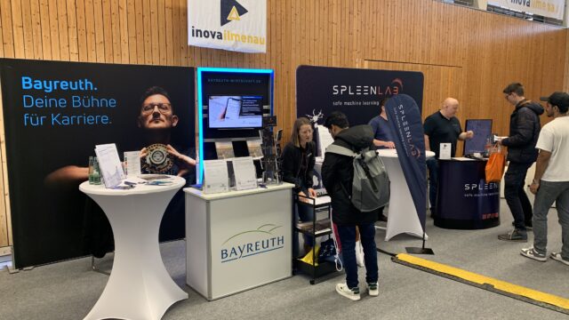 Eine Person steht an einem Messestand in einer Halle im Gespräch mit einer Frau. Im Hintergrund steht. Bayreuth. Deine Bühne für Karriere
